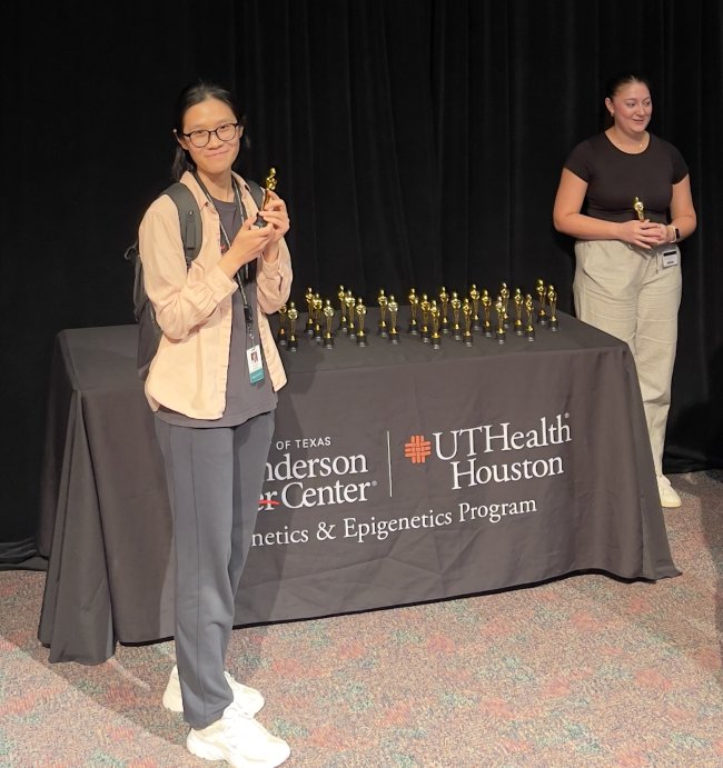 Student Lanxin Bei standing next to award table holding her award with one of the emcee's Renee Rubiano standing to the right of the table.