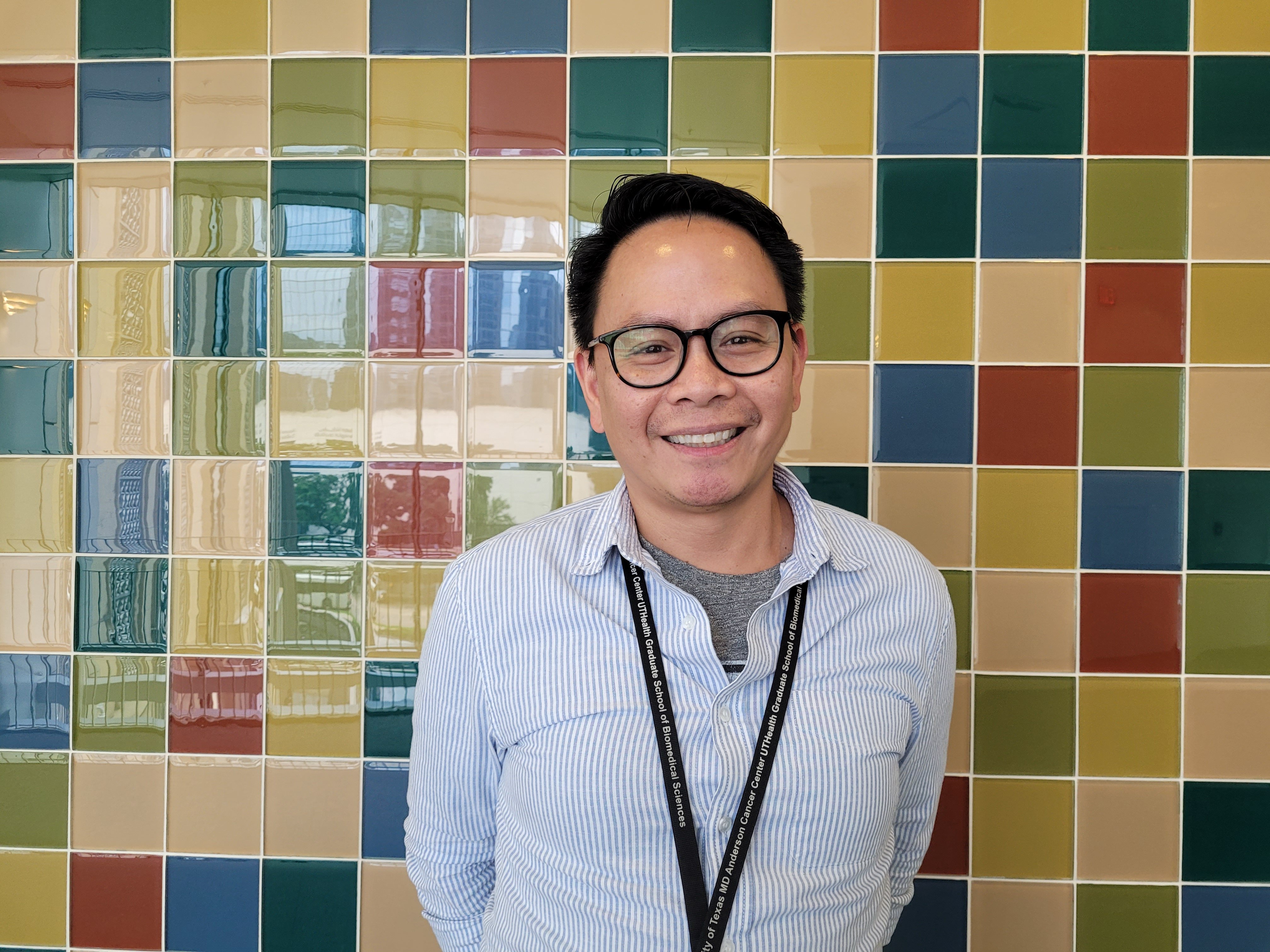 Photo of a man with black hair and glasses smiling in front of a multicolored tile wall
