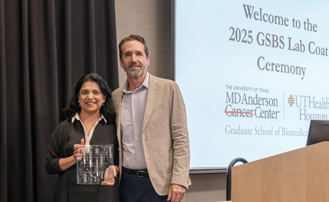 Swathi Arur, PhD holding a glass award beside Dean Alejandro Aballay at the 2025 GSBS Lab Coat Ceremony, with a projected slide in the background showing the MD Anderson Cancer Center and UTHealth Houston Graduate School of Biomedical Sciences logos