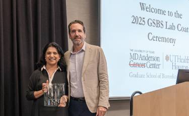 Swathi Arur, PhD holding a glass award beside Dean Alejandro Aballay at the 2025 GSBS Lab Coat Ceremony, with a projected slide in the background showing the MD Anderson Cancer Center and UTHealth Houston Graduate School of Biomedical Sciences logos