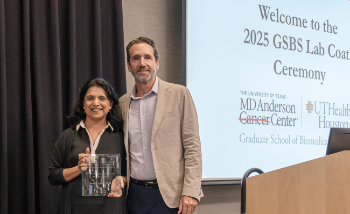 Swathi Arur, PhD holding a glass award beside Dean Alejandro Aballay at the 2025 GSBS Lab Coat Ceremony, with a projected slide in the background showing the MD Anderson Cancer Center and UTHealth Houston Graduate School of Biomedical Sciences logos