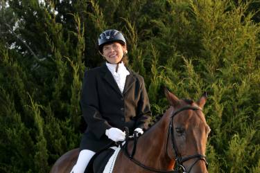 Karin Loftin smiles while seated on a brown horse, wearing formal equestrian attire and a riding helmet outdoors.
