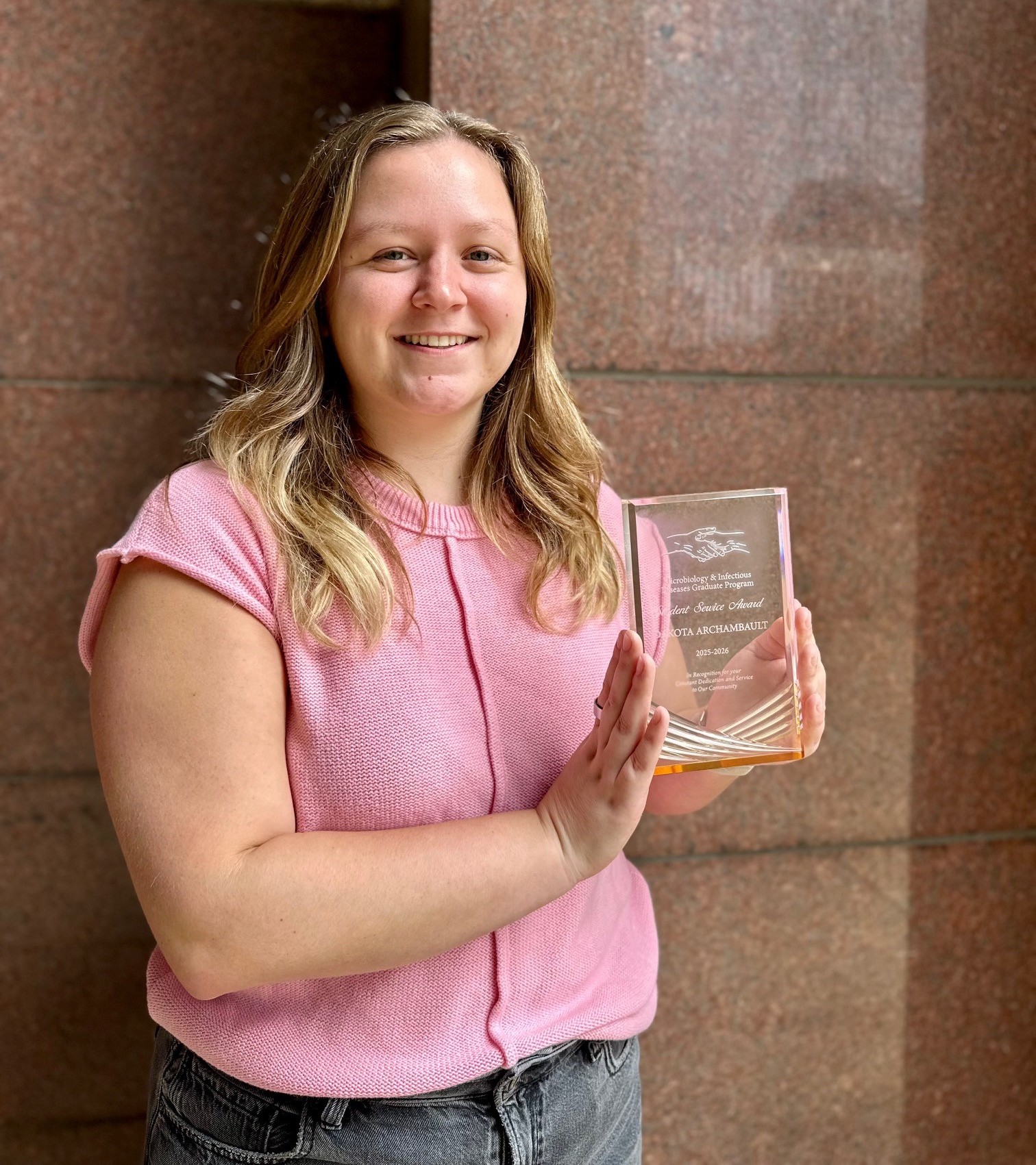 Woman in a pink top holding a service award plaque in front of a stone wall