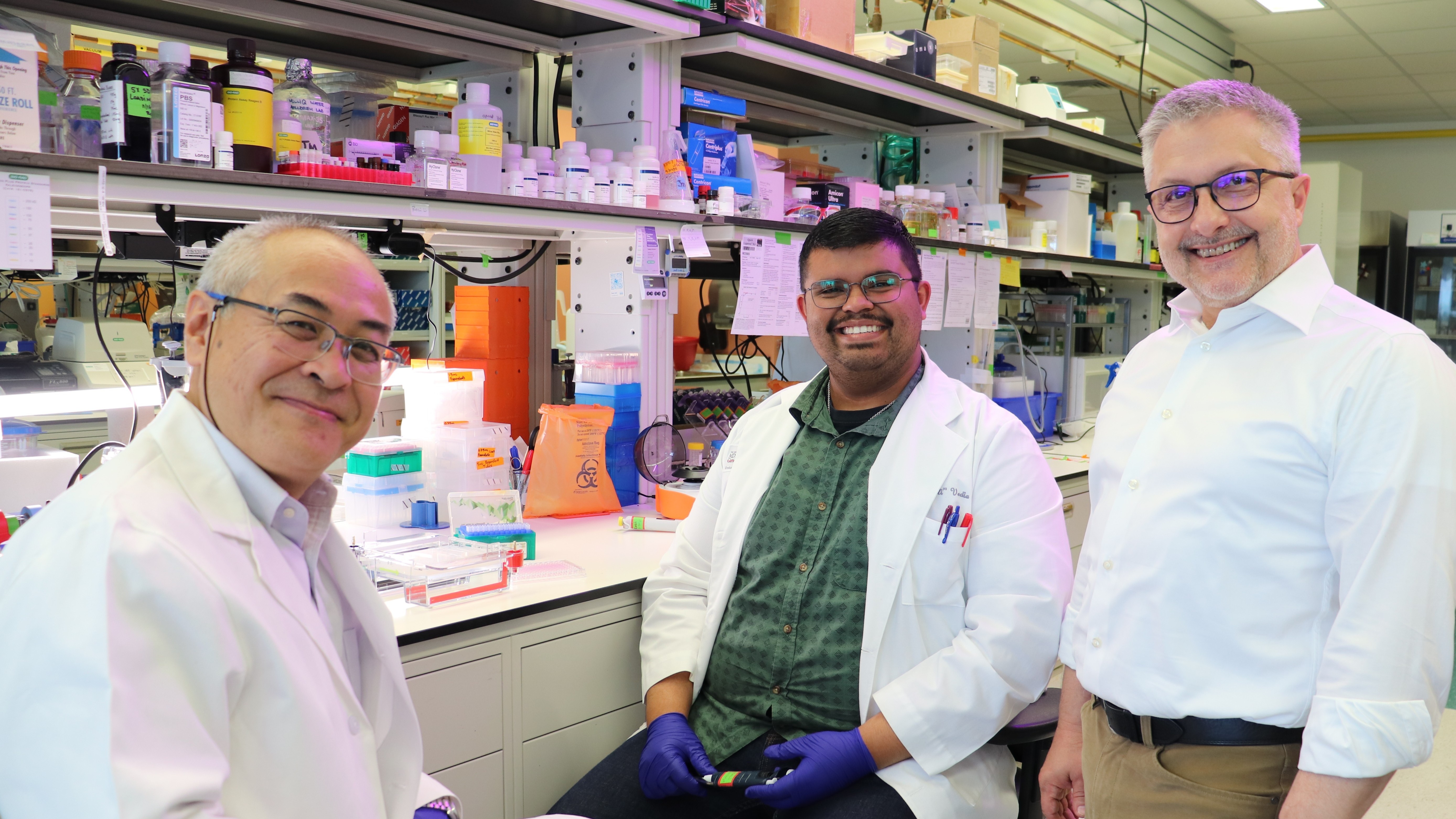 Three researchers in lab coats smiling in a laboratory, with equipment and chemical bottles on shelves behind them