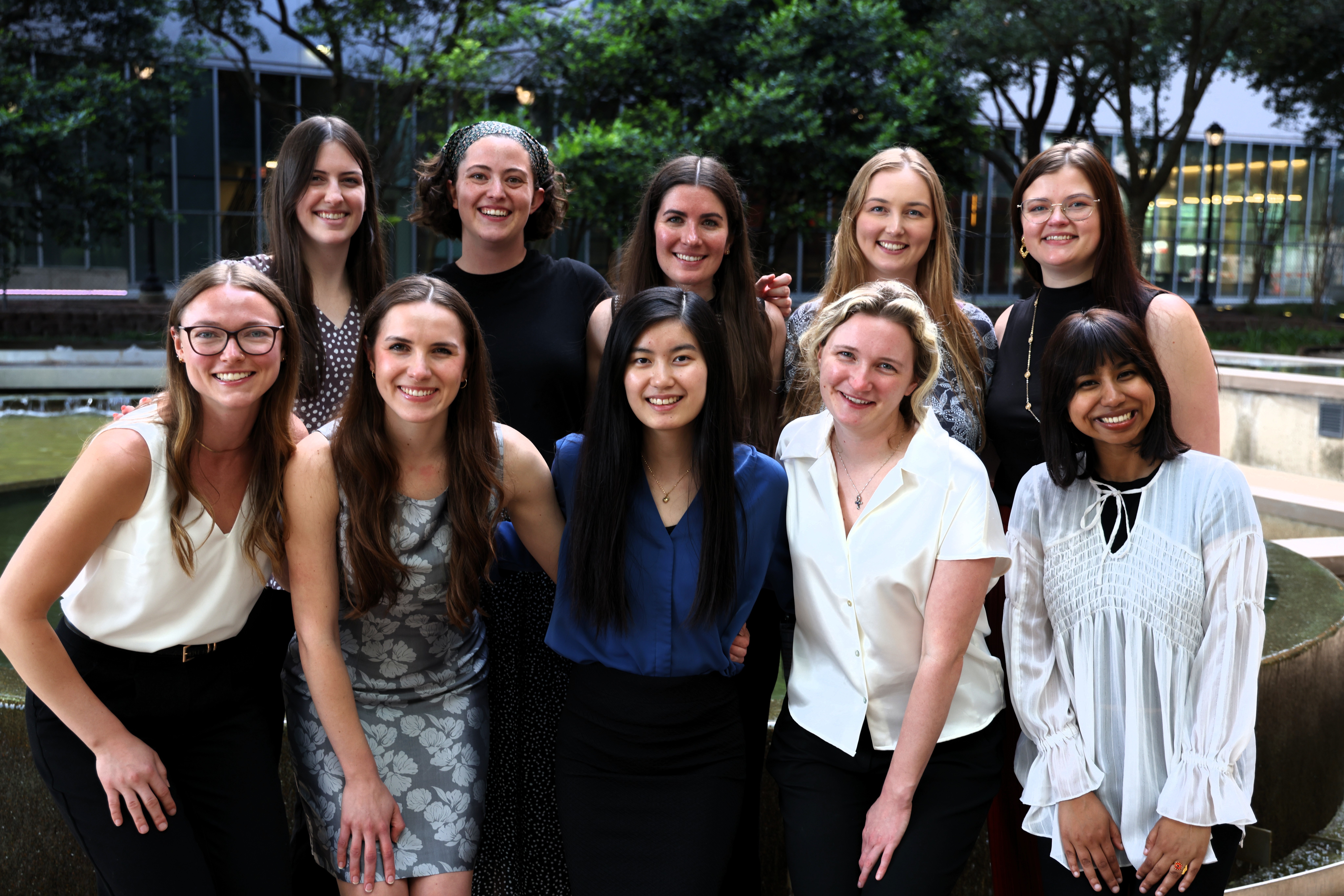 Group of women posing outdoors near a fountain and trees, smiling for a group photo