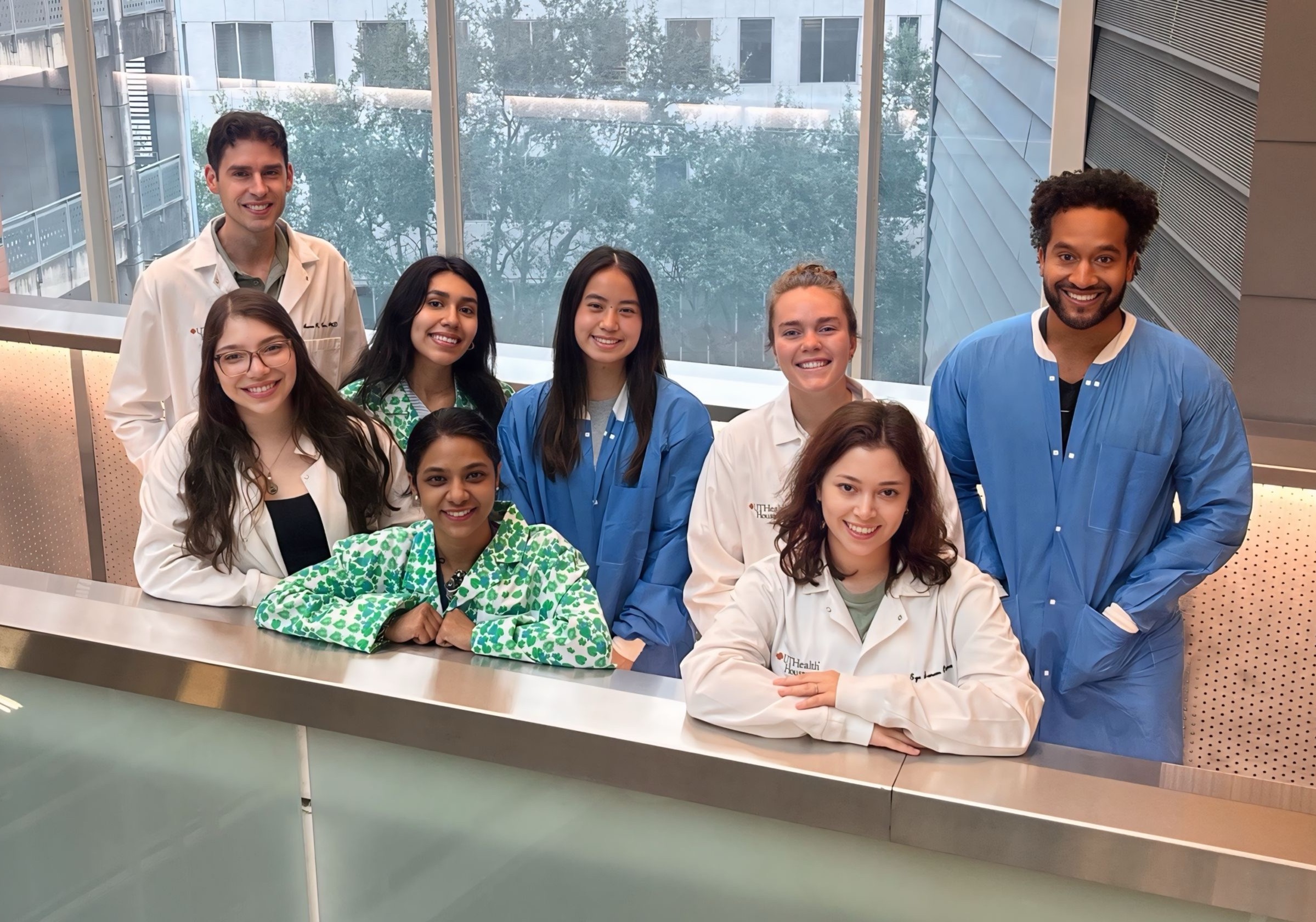 Group of students and staff in lab coats and scrubs posing indoors by a railing with campus buildings visible outside